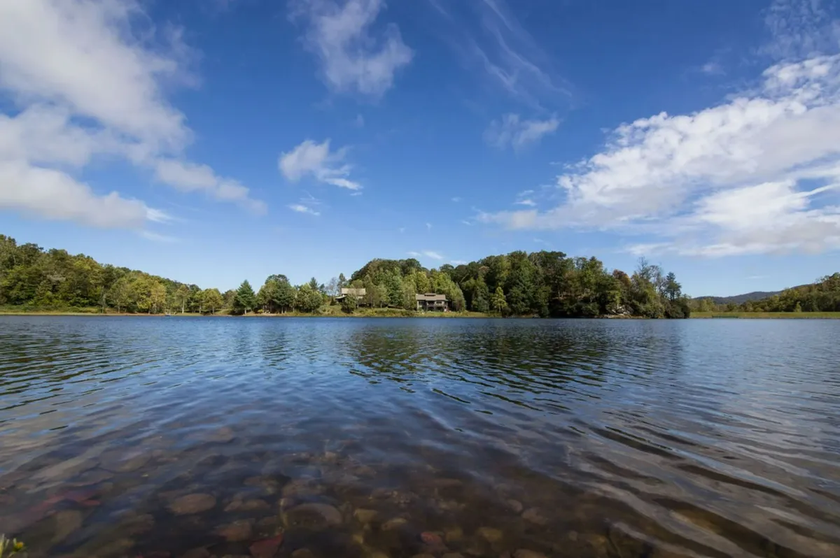 A tranquil view of the lake surrounded by lush greenery, with a distant house nestled along the shore. The calm water reflects the changing seasons, and the natural setting suggests opportunities for boating, fishing, bird watching, and tranquil relaxation.
