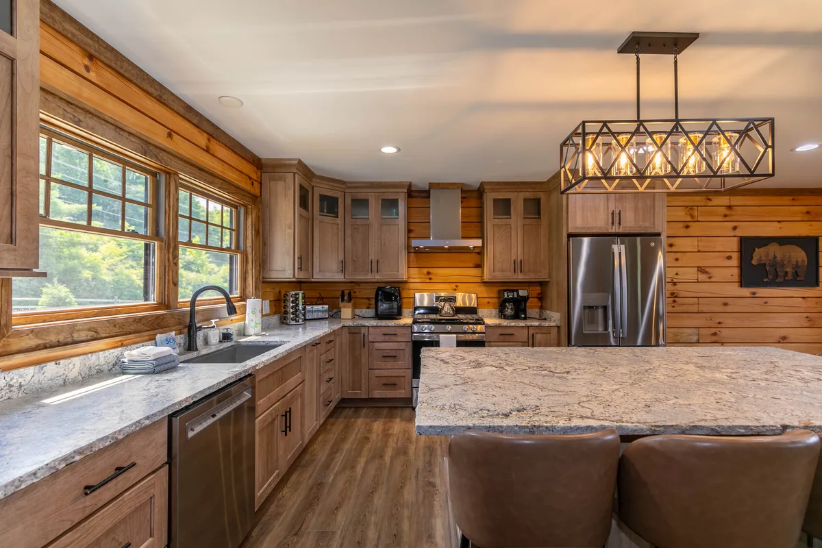 A gourmet kitchen with a large granite island, natural wood cabinetry, stylish chandelier lighting, and windows offering natural views. Leather barstools line the island, creating a space for both meal prep and socializing.