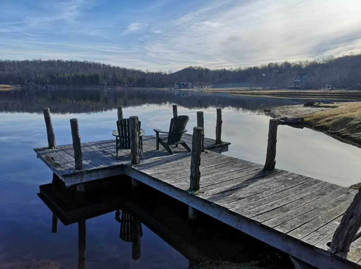 A wooden dock with adjoining chairs extending over calm water, framed by lush trees. The scene invites guests to fish, relax by the water, watch sunsets, or stargaze, with the dock serving as a serene vantage point for all-season enjoyment.