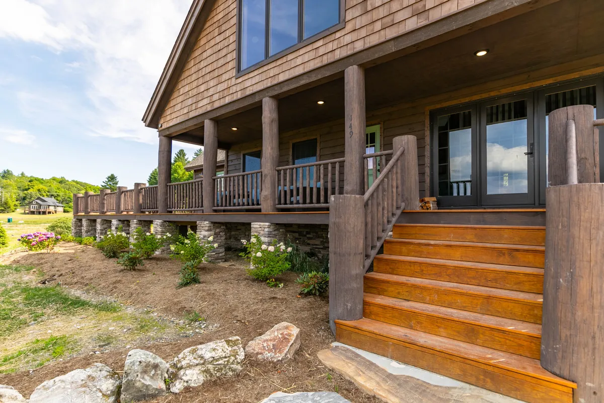 A welcoming wooden porch with stone foundation offers comfortable seating and space for socializing or quiet mornings. Surrounded by nature, vibrant flower beds line the entry, and the porch is positioned to capture sunlight and views, suggesting a perfect spot for relaxation or friendly gatherings.