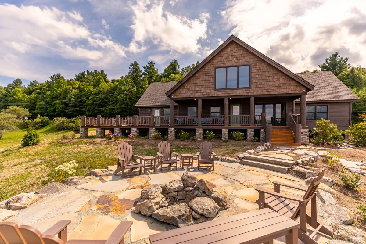 The image showcases the exterior of a brand new mountain home with a spacious wooden deck overlooking lush greenery. A stone fire pit sits ready for gatherings, while large windows hint at cozy interiors. The serene setting invites guests to relax outdoors, enjoy sunsets, and connect with nature.