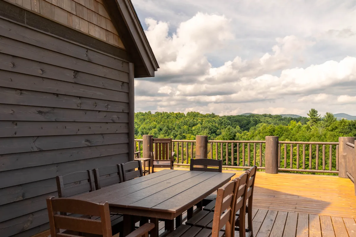 A spacious wooden dining table sits on an open-air deck, bordered by a log railing and framed by the surrounding forest. The setting is ideal for alfresco dining, with ample room for family and friends to gather amid serene woodland views.