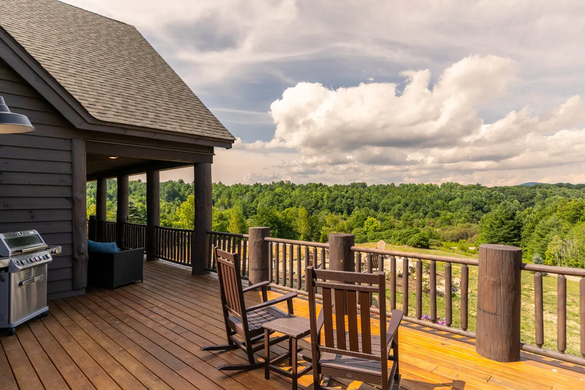 An expansive deck with wooden chairs and a grill, offering sweeping views of trees and hills beyond. This area provides a spacious, open-air environment ideal for gatherings, barbecues, and taking in the surrounding landscape.