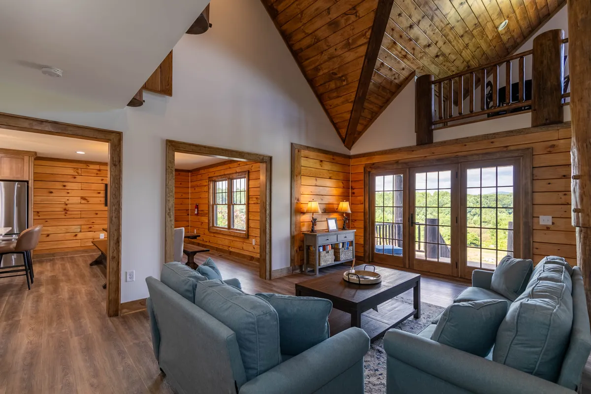 A spacious living area with a vaulted wooden ceiling and large windows that let in scenic natural light. The room features a comfortable sofa and warm wooden furniture, creating a welcoming space for family gatherings and relaxation.