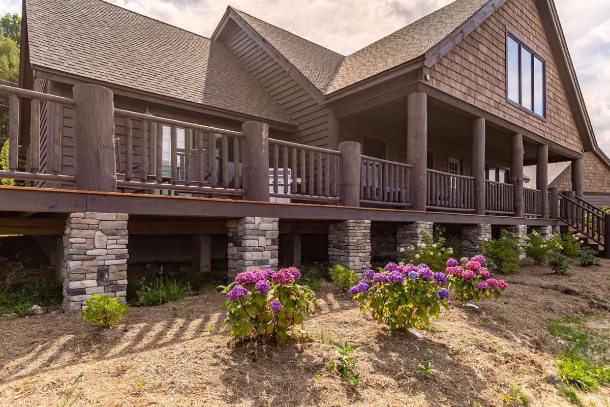This image features a welcoming wooden deck adorned with colorful hydrangea flowers, a sturdy stone foundation, and large windows that hint at the spaciousness inside. The deck flows seamlessly into a garden area, evoking a tranquil, nature-connected setting ideal for relaxing or hosting outdoor gatherings.