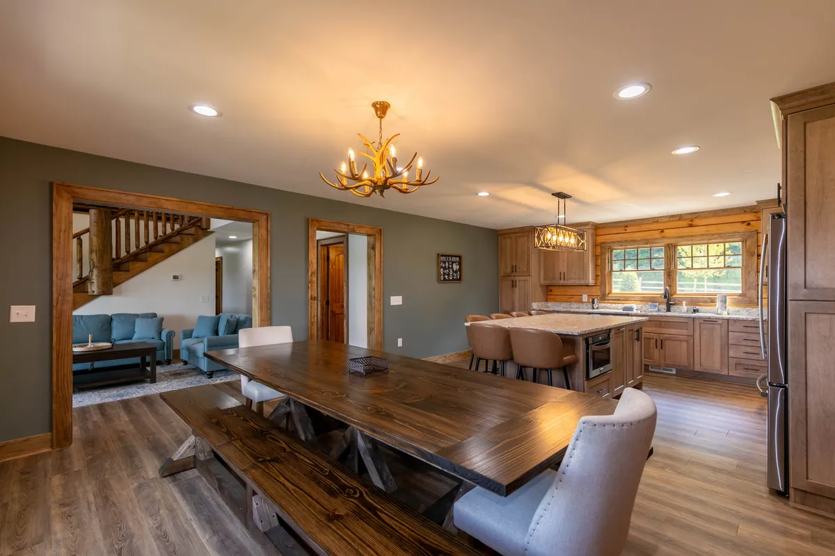 An open-concept dining and kitchen area featuring a long dining table, a granite-topped kitchen island with barstools, a modern chandelier, and a visible staircase. The arrangement encourages communal meals and lively gatherings.