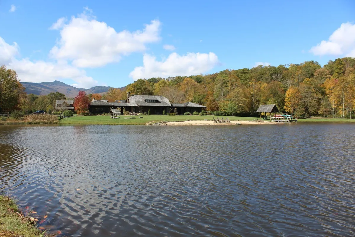 This wide exterior shot displays a large rustic building set beside a serene lake, with a small sandy beach, surrounding trees, and distant mountains creating a scenic backdrop. The landscape offers ample space for outdoor activities such as hiking, wildlife observation, and lakeside adventures.