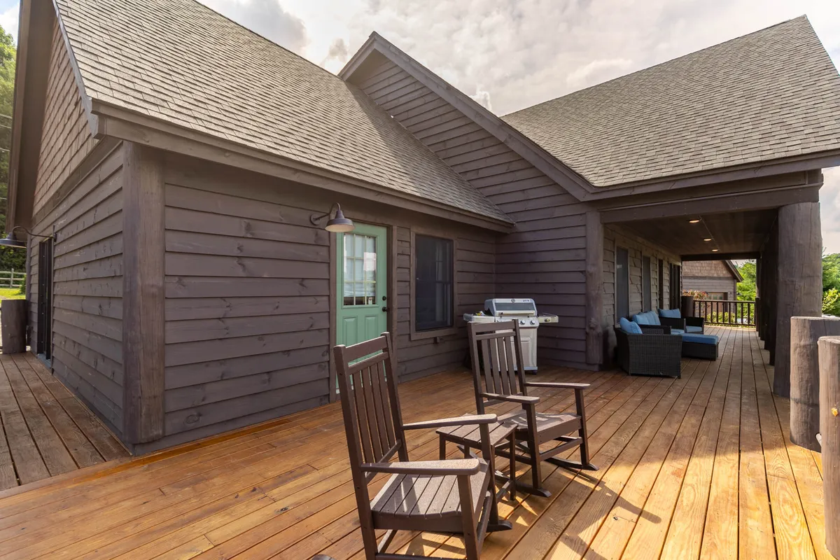 This image features a welcoming wooden deck equipped with classic rocking chairs, a green door leading inside, and an outdoor seating area with a grill. Surrounded by lush trees, the space invites relaxation and social gatherings with the outdoors just steps away.