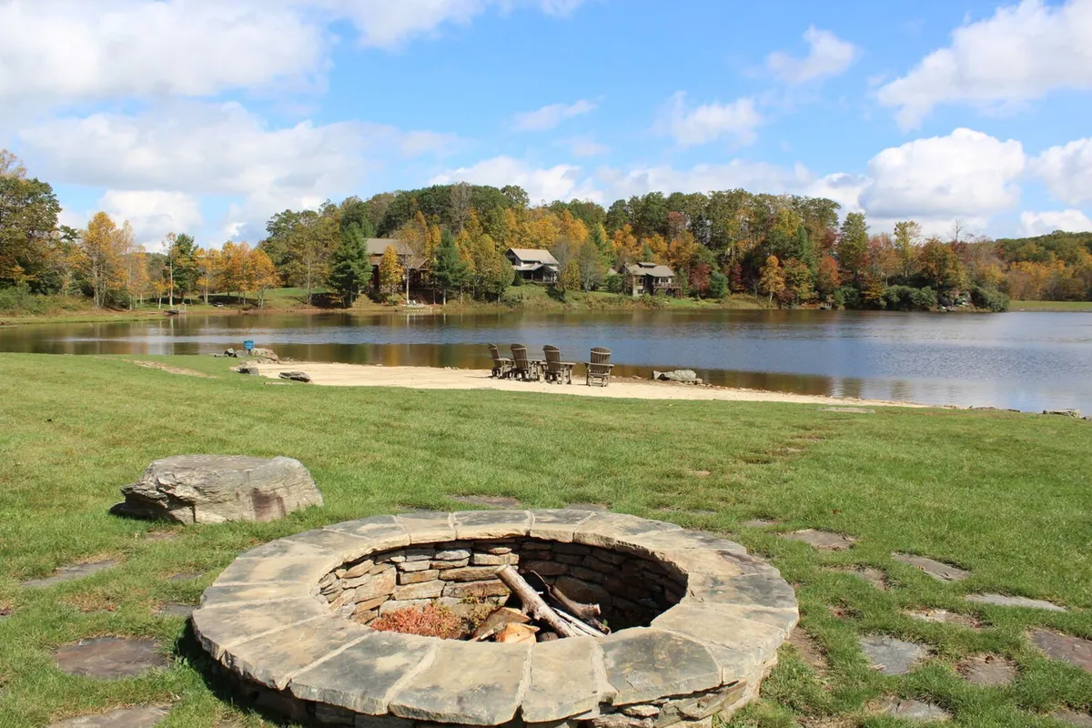 A stone fire pit with wooden chairs situated by a calm lake and framed by autumn foliage. The setting evokes warmth, camaraderie, and the joy of gathering around a crackling fire in nature's embrace.