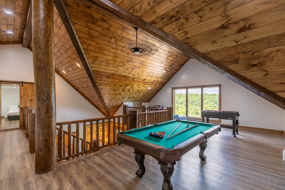 A bright, open loft featuring a pool table and foosball table under exposed wooden beams, with large windows inviting in natural light. Groups of people are seen engaging in both games, socializing, and enjoying the vibrant, playful atmosphere.