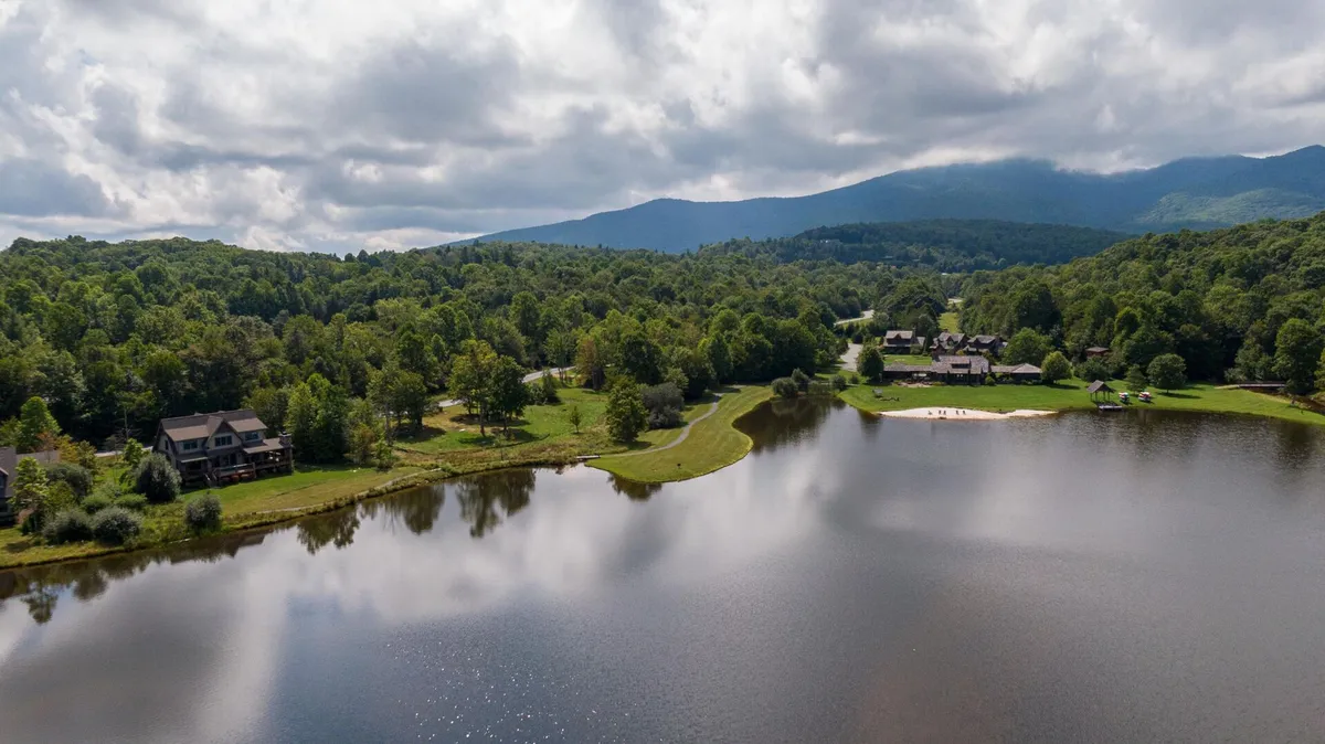 A sweeping view of a pristine lake bordered by mountains and trees, with houses dotting the shoreline. The scene captures the tranquility of the natural environment and evokes a sense of community amenities, like lakeside picnics or scenic strolls.
