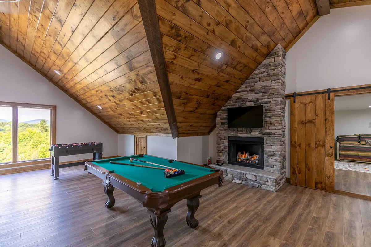 A stylish interior space with a pool table positioned near a striking stone fireplace and large windows. The room offers a cozy ambiance, ideal for both spirited games and relaxed evenings by the fire.