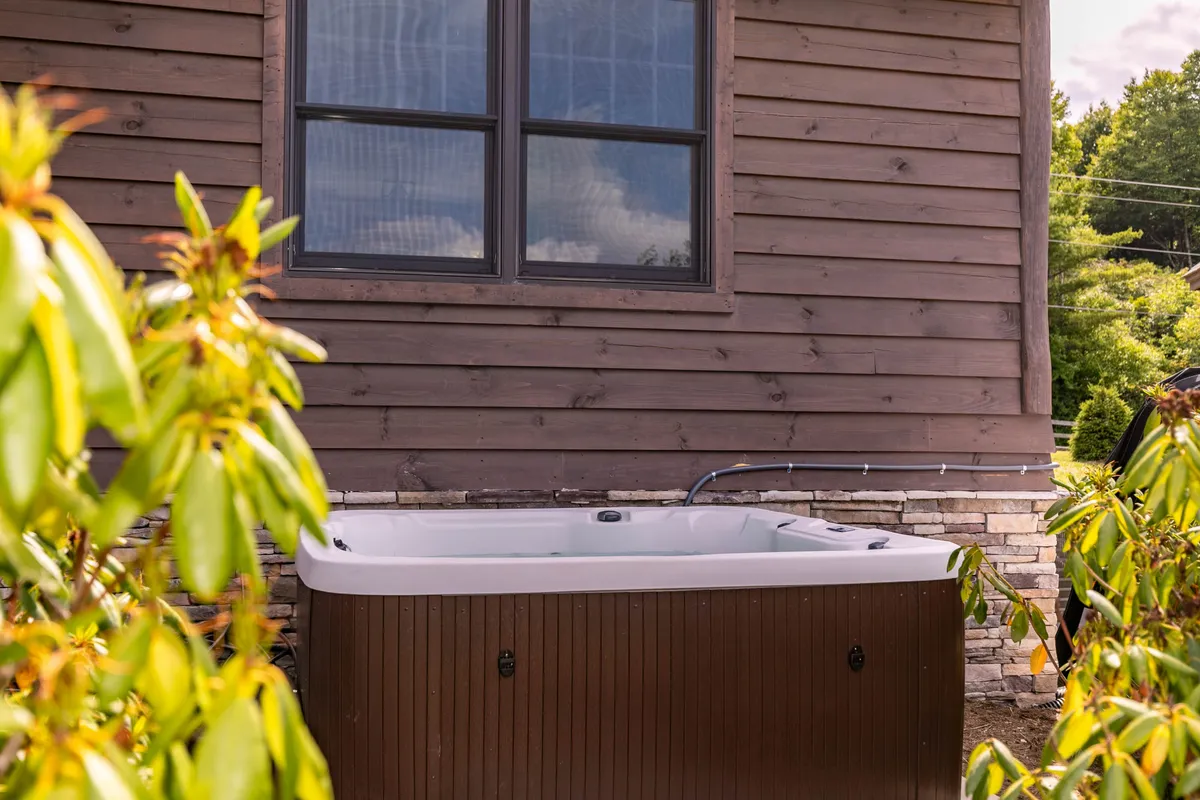 An inviting outdoor hot tub nestled beside the wooden exterior of the home, surrounded by lush foliage. The space radiates tranquility, perfect for stargazing or soothing soaks after a lively evening indoors.